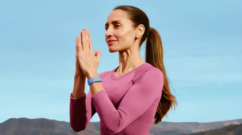 An individual dressed in light purple appears to be doing yoga outside while wearing a light blue Fitbit Air on their left wrist
