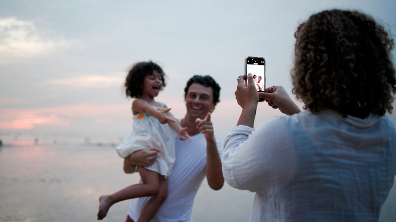 Mother captures a picture of father holding their daughter on a beach