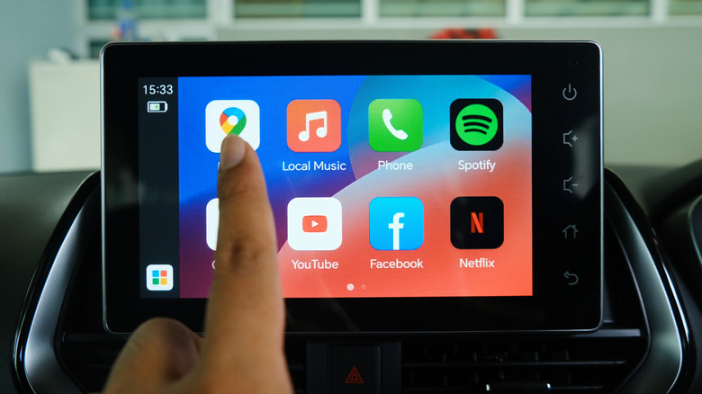 A finger pointing at the Google Maps icon as it appears on the Android Auto screen, several other icons can be seen. including Netflix, Spotify, and Facebook