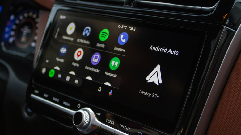 A black and white Android Auto icon appearing just below the same words on a vehicle's infotainment screen, the app drawer of Android Auto is to the right and shows a number of apps, including a telephone app and Spotify