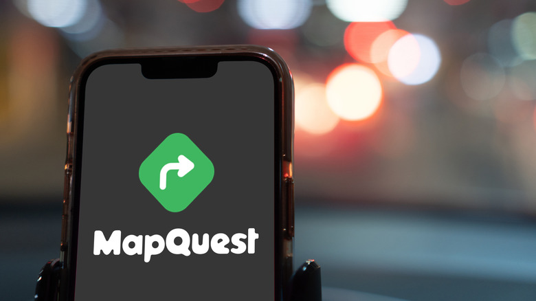A smartphone displaying the MapQuest logo as it sits in a cellphone holder inside a vehicle, the vehicle appears to be outdoors, but the background is too blurred to make it out fully
