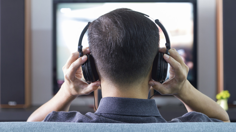 Taken from behind the user's head, an individual wearing a pair of black headphones faces a TV that is too blurred to discern what's going on, the user has both hands near each speaker as the headphones sit on their head
