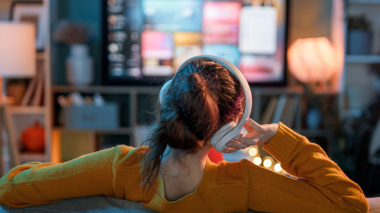 An individual in a yellow sweater is wearing a pair of white over-ear headphones as they watch TV. Shot from behind, the user has their right hand pressed against the earphones on their right ear