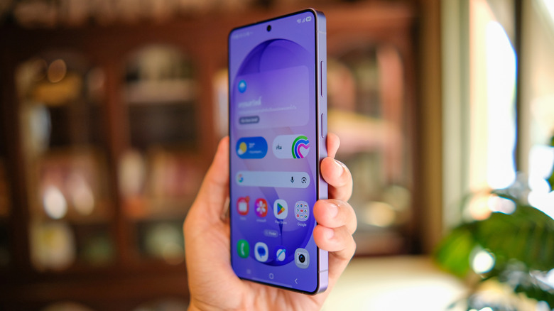A Samsung Galaxy S26 smartphone held in a user's left hand, though it's slightly tilted to display the Home screen at an angle, there appears to be a blurry set of shelves and a plant in the background