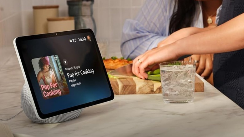 An Amazon Echo Show displaying a playlist about cooking, the device sits next to a counter while two individuals are cutting vegetables