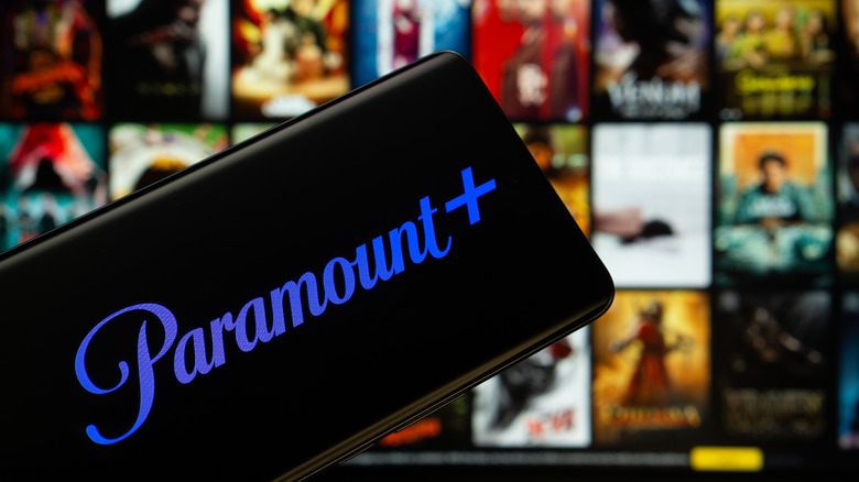 The Paramount+ logo is displayed in blue on a cellphone screen with a variety of movies and shows title cards are blurred in the background