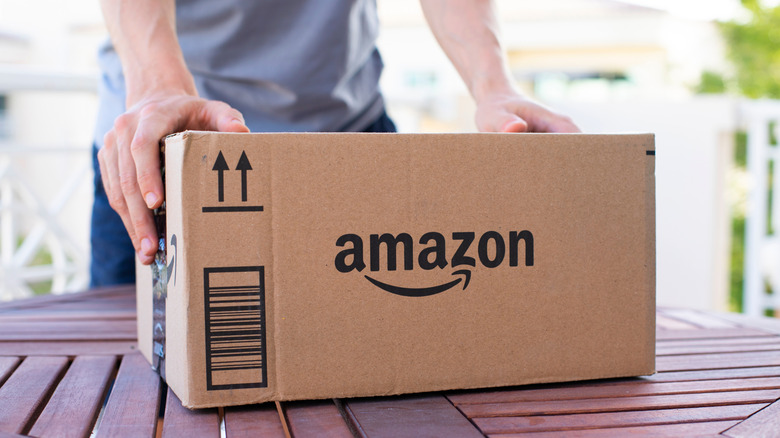 A closeup of an Amazon package as it sits on an outdoor table, an individual has both of their hands placed on the cardboard box, which is also prominently displaying the Amazon logo