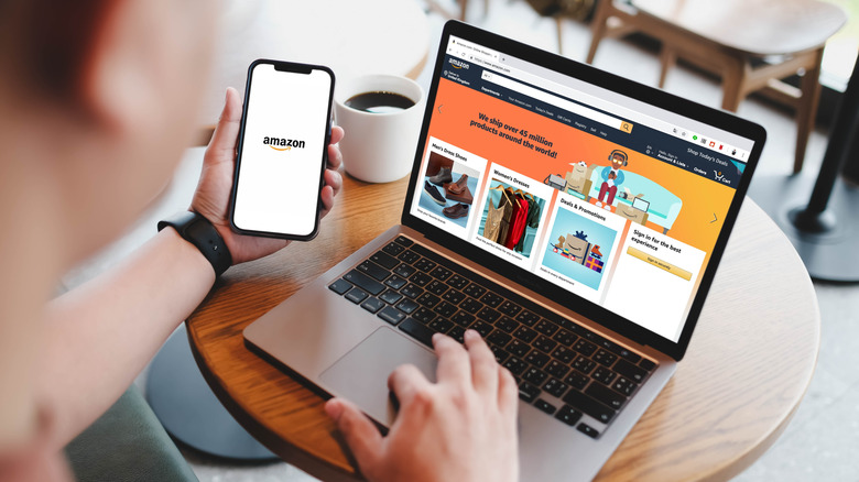 An individual holds a smartphone displaying the Amazon logo in their left hand as they navigate a silver laptop displaying the Amazon home page with their right