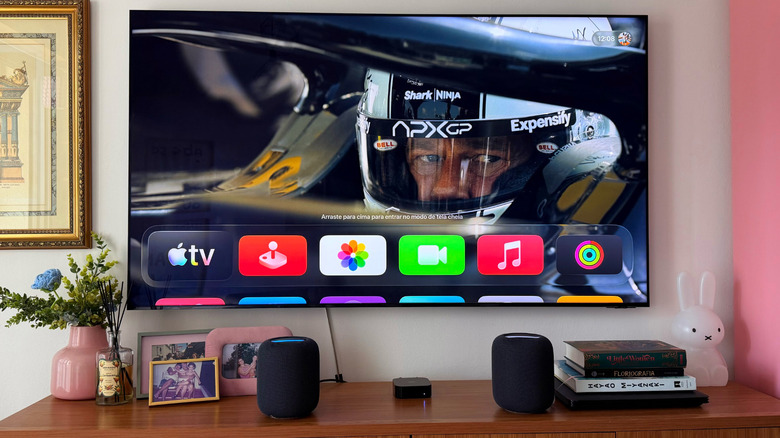 A smart TV displaying the Home Screen of an Apple TV. There are also a pair of HomePod speakers, a few books, frames, and a plant.