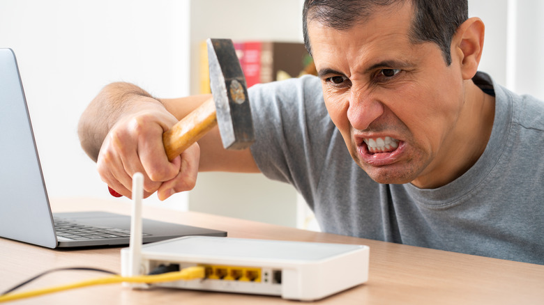 An angry man takes a hammer to his internet Wi-Fi router besides his open laptop.