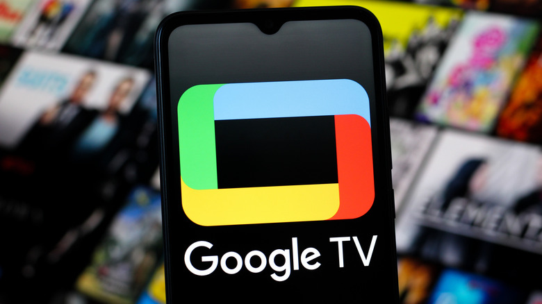 A smartphone displaying the Google TV logo, the device is in front of a blurred background that appears to be a movie selection screen for a streaming service or a smart TV