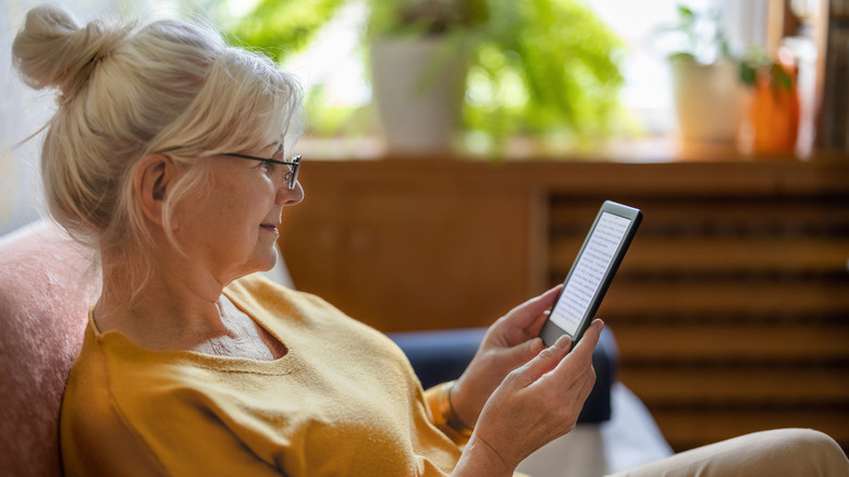 An older woman with glasses looks smilingly on her Kindle as she reads it on her couch