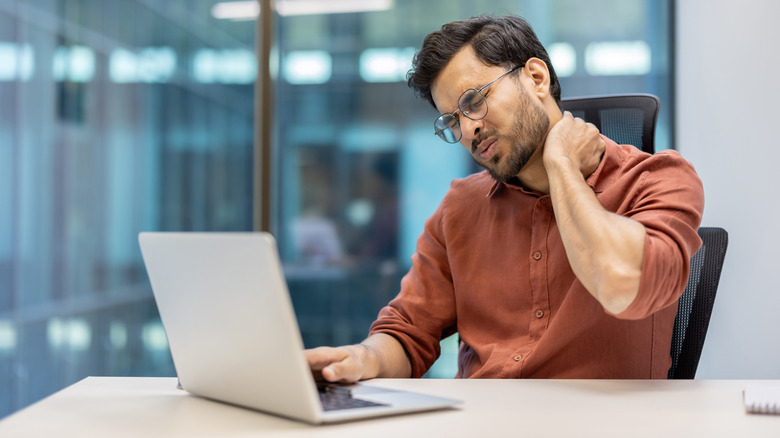 Man in an office looking down at a laptop while holding his neck in pain