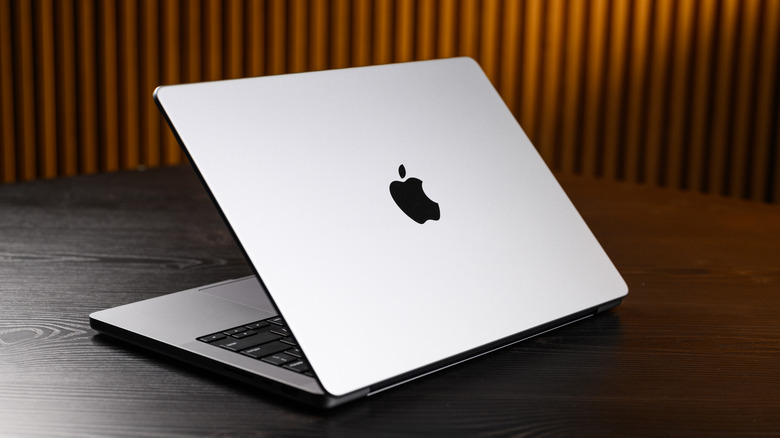 A grey M2 MacBook Pro sitting on a darkly-colored table, the screen is half open, and the device's back is facing the camera