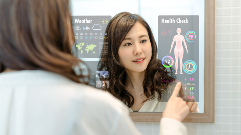 A brown-haired woman smiling as she places her finger on a smart mirror displaying health and weather data.