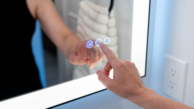 A close up of a person's hand tapping the light button on a bathroom smart mirror.
