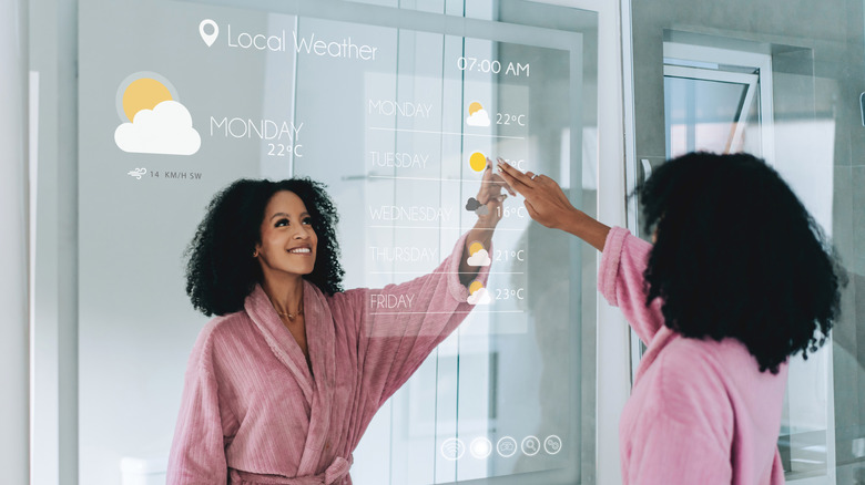 A woman in a pink robe checking the weather forecast using a smart mirror in a bathroom.