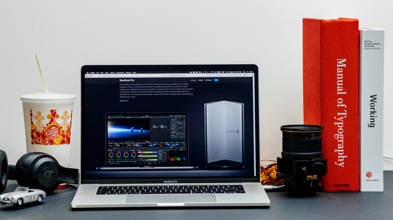 A silver 2018 Apple MacBook Pro on a surface flanked by books, a camera lens, Beats headphones, and a Burger King cup.