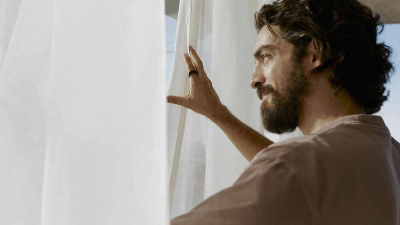 A man pulling the curtains to look through a window while wearing an Oura Ring.