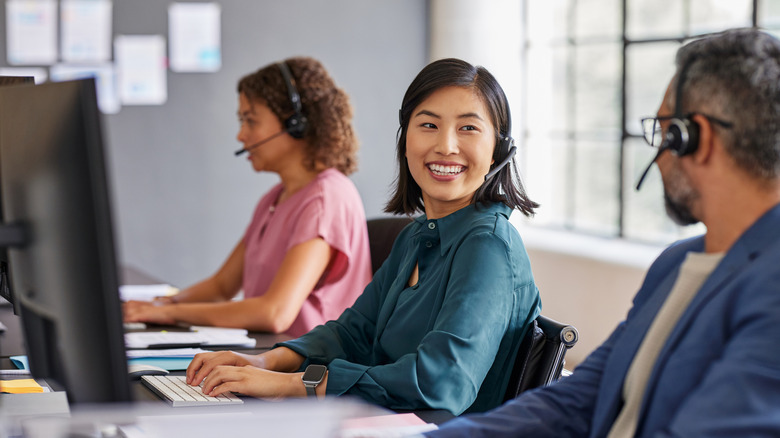 A group of smiling call center employees talk with one another while working at their desks.