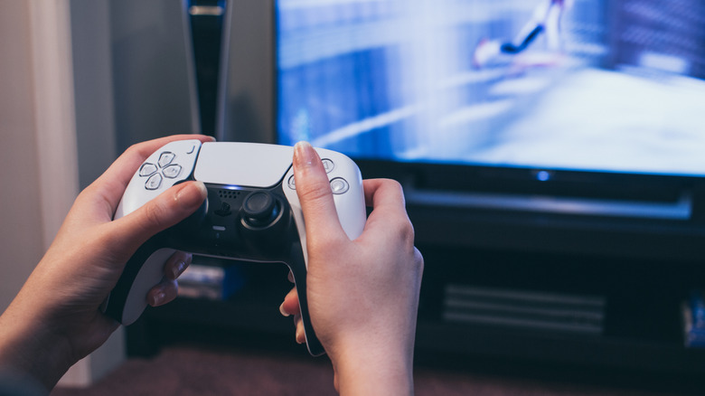 A gamer holds a PS5 controller with a gaming console and TV in the background