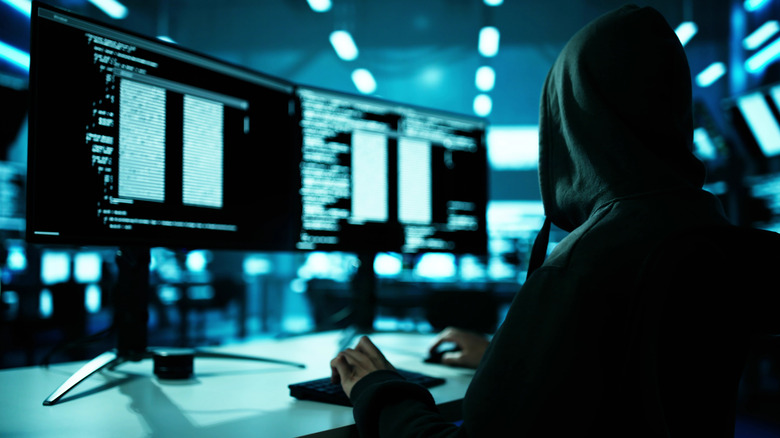 A hacker wearing a hood while sitting at a desk and looking at code on two computer monitors