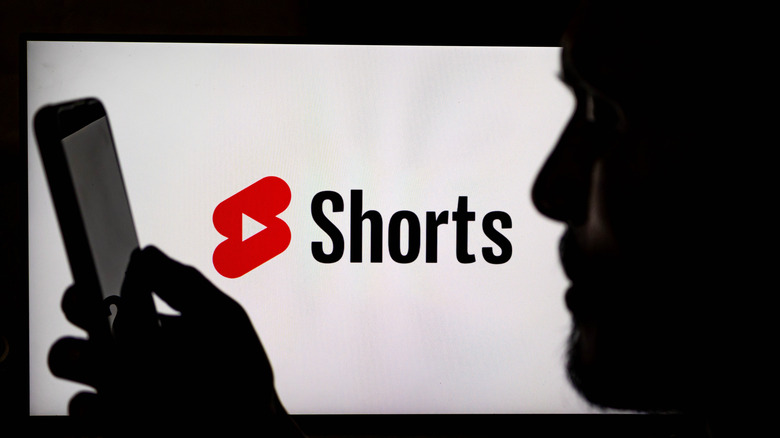 Silhouette of a person watching YouTube shorts on their smartphone with logo behind.