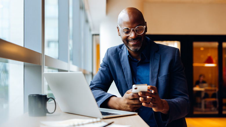 A man in a shop smiling while holding his phone in front of a laptop.