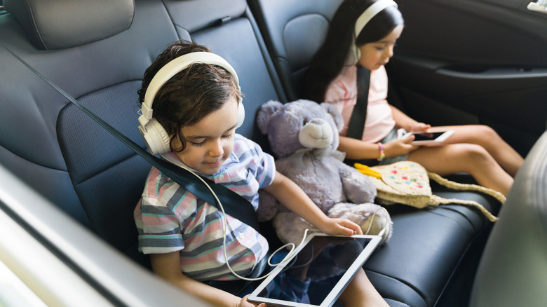 Two kids in the backseat of a car wearing headphones and on devices.