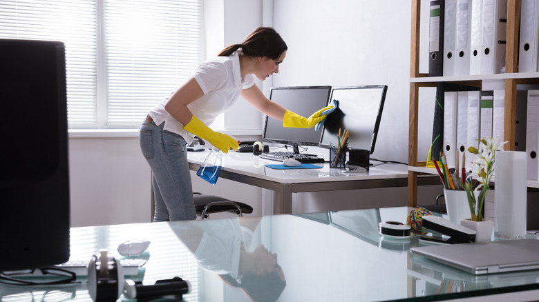 Woman cleaning a computer desk and monitors in an office setting.