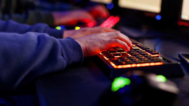 Close up of a person's hands on an RGB keyboard in front of computer monitors.